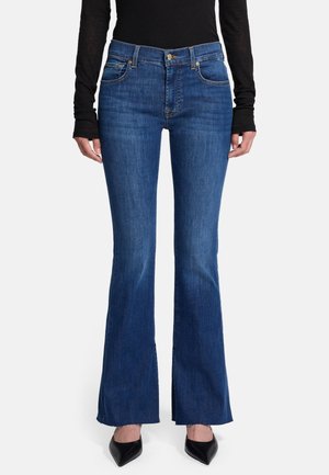 Flared blauwe jeans gemaakt van denim met een donkere wassing, voorzien van voorzakken, een ritssluiting en splitjes bij de enkels; gedragen met zwarte puntige schoenen.