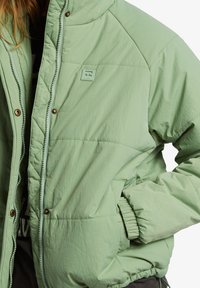 Giubbotto puffer verde chiaro con design trapuntato, bottoni a pressione e polsini elasticizzati. Presenta un'etichetta con il logo sul petto e tasche laterali.