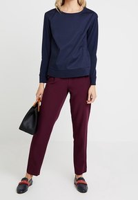 Marineblaues Langarmoberteil mit rundem Ausschnitt, kastanienrote gerade geschnittene Hose, schwarze Handtasche und dunkle Schuhe mit rotem Akzent.
