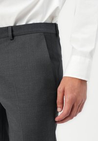 Grå dressbukser laget av glatt stoff, med slim fit, sidelommer og en ren midje. En hvit skjortekant er delvis synlig.