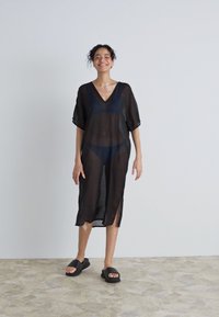 Black, sheer kimono-style cover-up with a V-neck, short sleeves, side slits, and a loose fit, worn over dark swimwear. Flat black sandals.