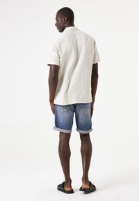 Lichtbeige shirt met korte mouwen, gestructureerde stof, gecombineerd met opgerolde denimshorts en zwarte sandalen, gezien vanuit de achterkant.