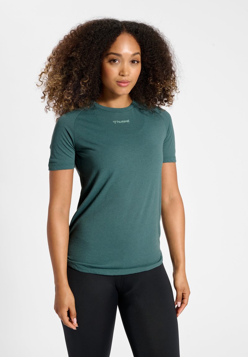 Femme aux cheveux bouclés portant un t-shirt de sport vert à manches courtes et un legging noir, debout sur un fond blanc.