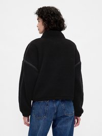 Μαύρο φλις μπουφάν με ψηλό γιακά, υπερμεγέθη σχεδιασμό και υφή επιφάνειας. Συνδυάζεται με μπλε τζιν ψηλόμεσο, εμφανιζόμενο από πίσω.