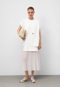 Un dessus blanc oversized avec un col rond, associé à une jupe plissée blanche et des baskets beiges. Tient un petit sac beige avec une bandoulière.