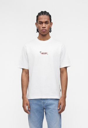 Νέος άνδρας με πλεξούδες, που φοράει λευκό μπλουζάκι με μικρό λογότυπο "REPLAY" και ανοιχτό μπλε τζιν, στέκεται μπροστά σε απλό φόντο.