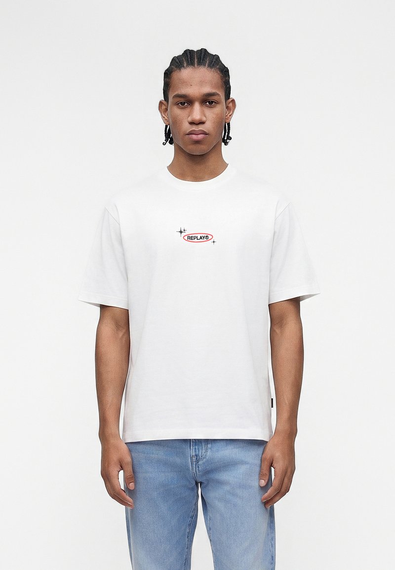 Νέος άνδρας με πλεξούδες, που φοράει λευκό μπλουζάκι με μικρό λογότυπο "REPLAY" και ανοιχτό μπλε τζιν, στέκεται μπροστά σε απλό φόντο.