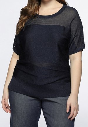Vrouw draagt een donkerblauw gebreid T-shirt met korte mouwen en een doorschijnend bovenpaneel, en donkerblauwe spijkerbroek, staand met ontspannen handen.