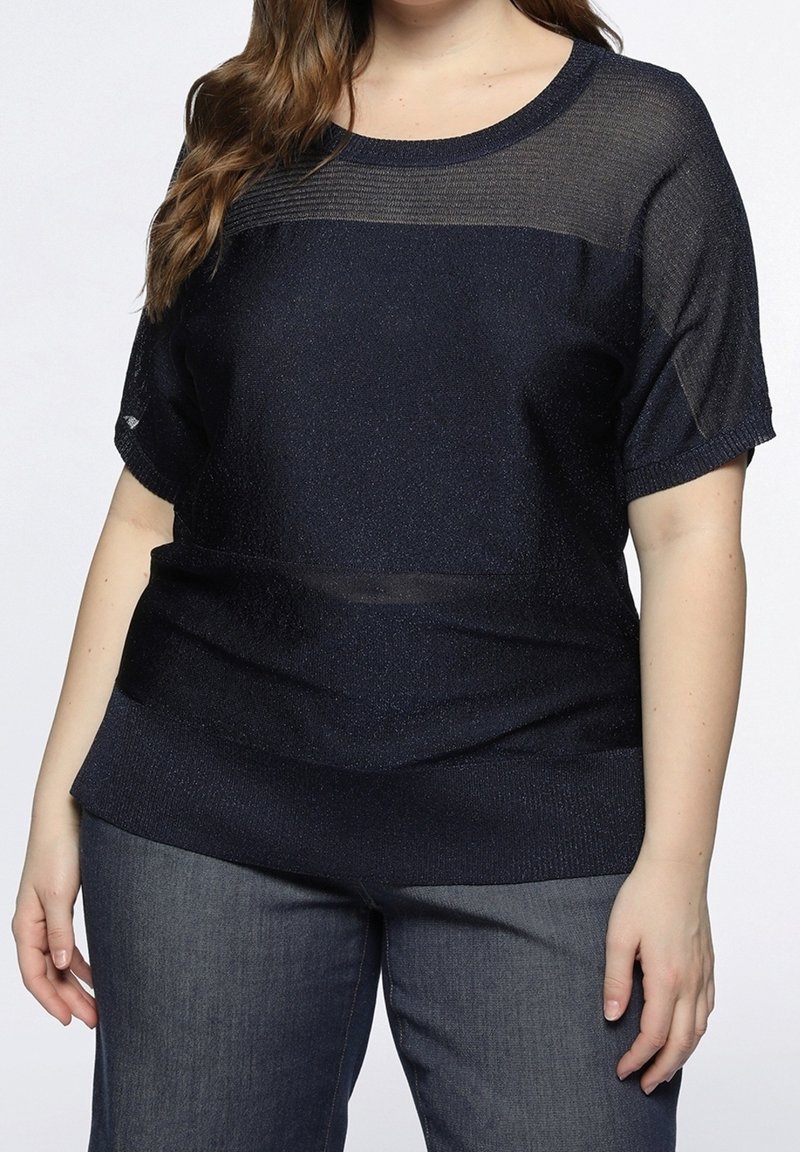 Femme portant un haut en maille bleu foncé à manches courtes avec un panneau supérieur transparent et un jean bleu foncé, debout avec les mains détendues.