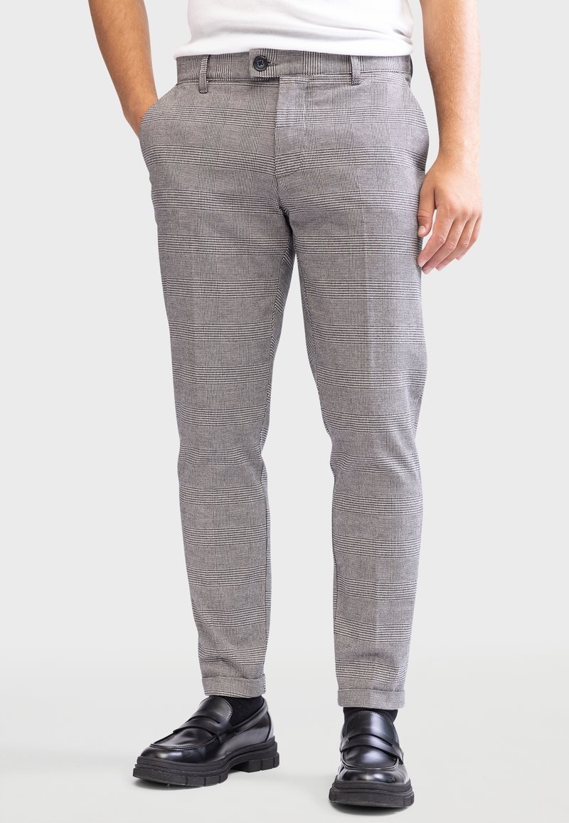 Homme portant un pantalon de costume gris à carreaux, des mocassins en cuir noir et une chemise blanche rentrée, debout avec une main dans la poche.