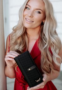 Black quilted wallet with a gold snap closure and logo detail, held by a person. The wallet features a smooth texture and rectangular shape.