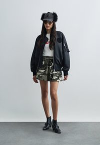 Veste bomber noire avec grandes poches, chemise blanche avec emblème rouge, jupe camouflage et bottines noires. Le mannequin porte des lunettes de soleil et un chapeau en fourrure.