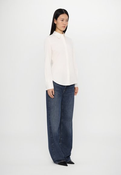 White button-up shirt with a collar, paired with wide-legged dark blue jeans and black pointed-toe shoes. Smooth fabric texture, clean lines.