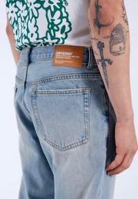 Ljusblå denimjeans med en blekt tvätt, som har en brun läderlapp på baksidan och två bakfickor med sömnadsdetaljer.