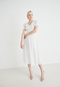 Valge midi kleit lühikeste lendlevate varrukatega, volditud bodidetailide ja keha rõhutatuna. Kombineeritud hõbedaste kõrgete kontsadega kingadega, millel on paelad.