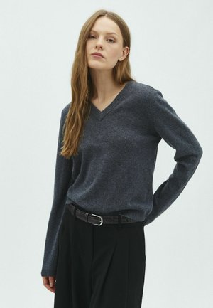 Femme aux longs cheveux bruns portant un pull gris foncé à col en V et un pantalon noir avec une ceinture noire, debout devant un fond uni.
