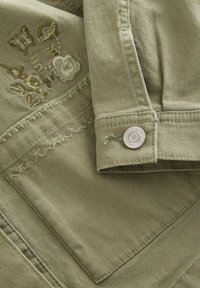 Khaki skjorta med blommönster i jordnära färger. Detaljerad sömnad på cuffs och ficka. Rund knapp med en subtil logo.