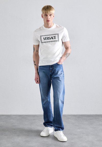 Tricou alb din bumbac cu logo-ul negru "VERSACE", asortat cu blugi din denim bleu deschis și adidași albi. Design simplu, croi lejer.