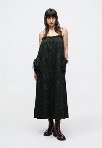 Μαύρο αμάνικο φόρεμα με υφή υφάσματος, σε σχήμα Α και με κλείσιμο με κουμπιά. Διαθέτει λεπτομέρειες με κορδόνια στα μανίκια. Φοριέται με κόκκινα γυαλιστερά μποτάκια.