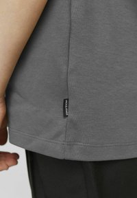 T-shirt grigio in cotone con orlo dritto, etichetta con marchio sulla cucitura laterale e maniche corte, mostrato accanto a pantaloni neri.