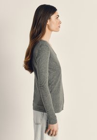 Ung kvinna med långt brunt hår som bär en grå långärmad tröja och ljusa jeans, stående i profil mot en enfärgad bakgrund.