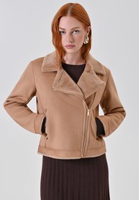 Giacca in finta suede color tan con colletto in pelliccia, chiusura con zip e due tasche laterali. Il design presenta una vestibilità sagomata e una texture morbida.