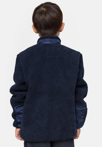 Enfant aux cheveux courts et foncés portant une veste polaire texturée bleu marine, vu de dos sur un fond clair uni.