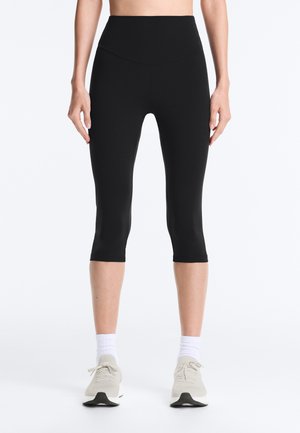 Femeie purtând colanți negri scurți, cu talie înaltă, șosete albe și pantofi sport bej deschis, stând pe un fundal alb simplu.