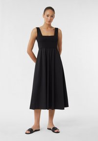 Schwarzes Midi-Kleid mit quadratischem Ausschnitt und tailliertem Oberteil, fließendem Rock und weicher Textur. Getragen mit flachen schwarzen Sandalen.