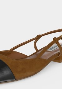 Barna velúr lapos szandál fekete bőr orrrésszel, vékony bokapánttal és kis négyzet alakú sarokkal fehér háttéren.