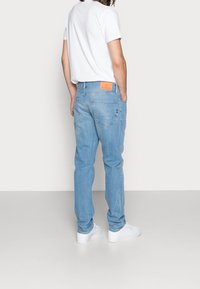 Man i ljusblå denimjeans med brun läderlogo och vita sneakers, stående med händerna i fickorna och vänd bort.