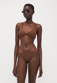 Brown textured bikini with a twist-front top and matching low-rise bottoms. Model wears black angular sunglasses. Simple and minimalist design.