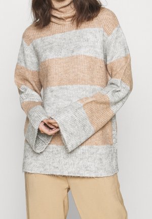 Pull à col roulé en maille côtelée ample avec larges rayures horizontales beige et gris clair, porté avec un pantalon beige, mains jointes devant.