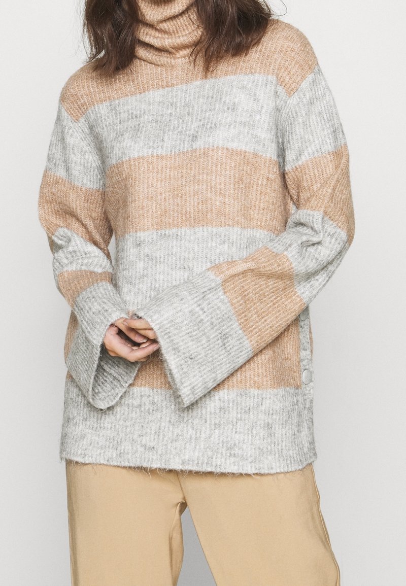 Pull à col roulé en maille côtelée ample avec larges rayures horizontales beige et gris clair, porté avec un pantalon beige, mains jointes devant.