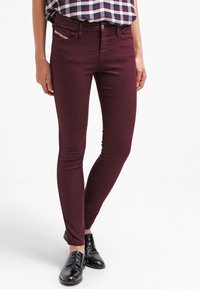 Person i vinröda skinny jeans, en rutig skjorta instoppad och svarta läderskor med snörning, står på en vit bakgrund.