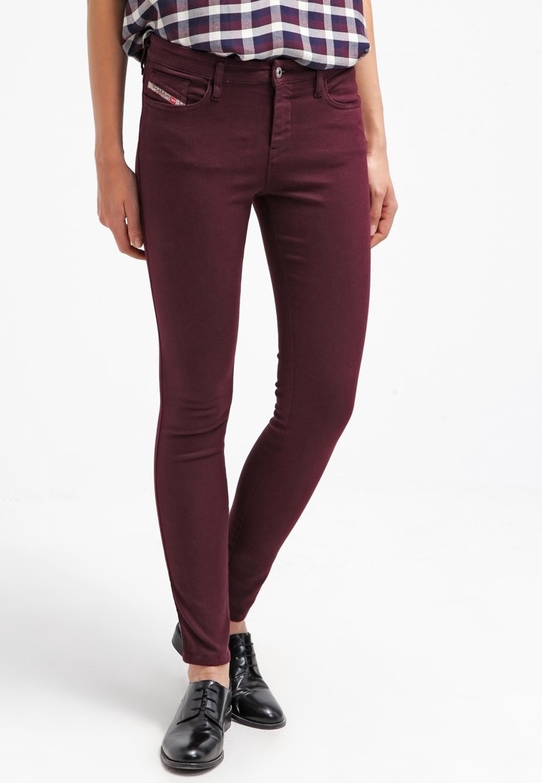 Person i vinröda skinny jeans, en rutig skjorta instoppad och svarta läderskor med snörning, står på en vit bakgrund.