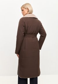 Brun polstret frakke med en beige faux fur krave, bælte i taljen og sidelommer. Har lodrette syninger og en knælang design.