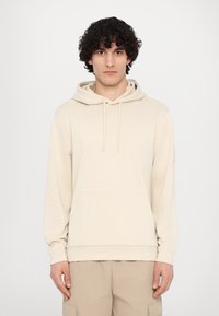 Felpa beige realizzata in tessuto morbido, dotata di tasca frontale, cappuccio con coulisse e polsini a costine. Il modello è in piedi contro uno sfondo neutro.