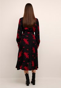 Robe noire à motifs floraux rouges, manches longues et ceinture. Tissu lisse avec une silhouette fluide, associée à des bottes à talons noires.