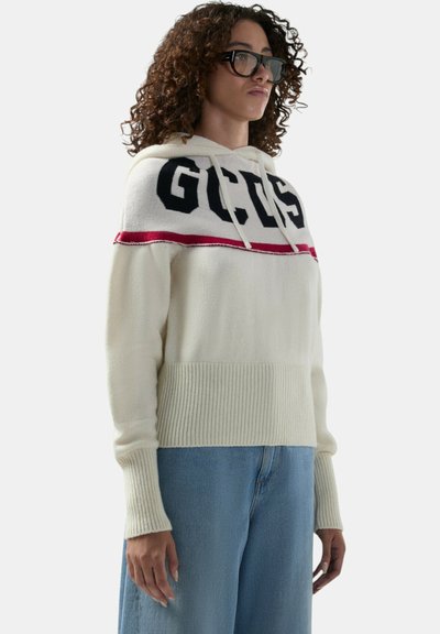 Persona che indossa occhiali neri oversize, una felpa con cappuccio crema lavorata a maglia con la scritta "GCDS" e jeans azzurri, in piedi contro uno sfondo neutro.