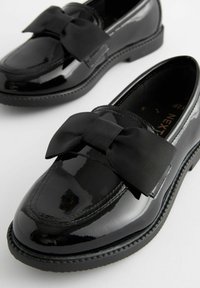 Mocassins en cuir verni noir avec un grand nœud en ruban noir à l'avant, avec une finition lisse et un détail texturé sur la pointe.