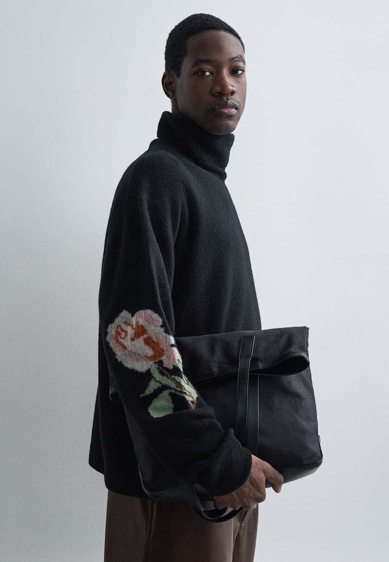 Bărbat îmbrăcat cu un pulover negru cu guler înalt, având un model floral pe manșon, ține o geantă mare neagră, stând în fața unui fundal simplu deschis.