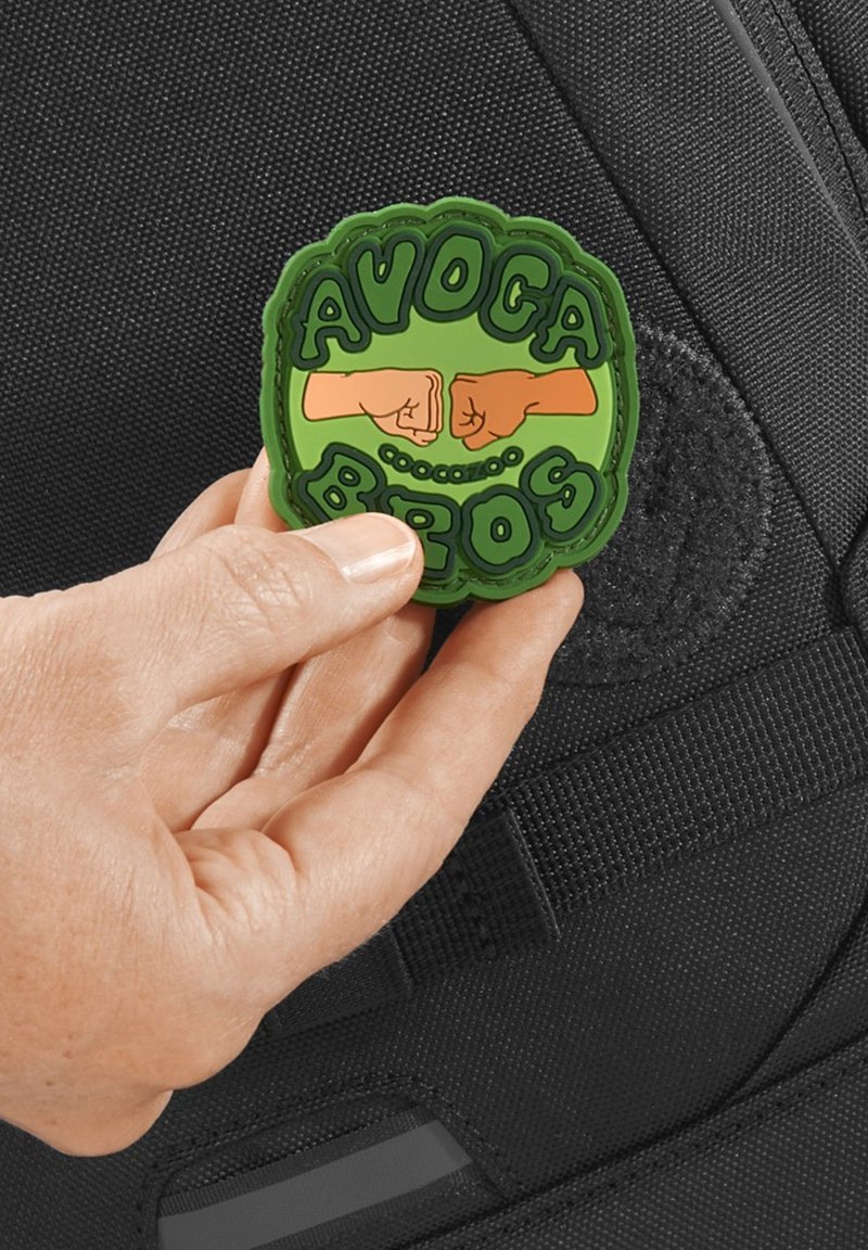 Green rubber patch featuring "AVOCA BROS" in raised letters with two fists bumping, held against a textured black bag.