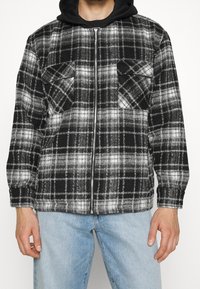 Black and white plaid shirt jacket with a zip front, two chest pockets, and a regular fit, paired with light blue jeans.