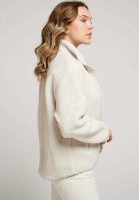 Veste en polaire douce couleur crème, avec col, fermeture éclair à l'avant et coupe décontractée. Présente des coutures visibles et des poches latérales.