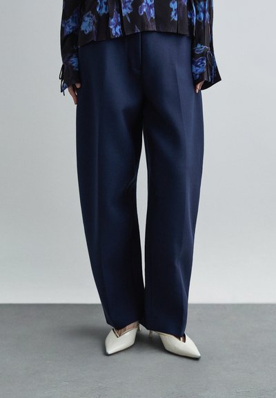 Pantalon large bleu marine en tissu lisse, porté avec des escarpins blancs à bout pointu. Motif floral visible sur le haut. Fond neutre.