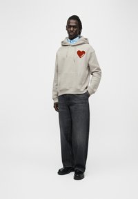 Sweat-shirt à capuche gris avec un graphique de cœur rouge, poche avant, associé à un jean en denim foncé à coupe ample et des chaussures noires. Fond uni.