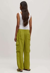 Groene, gekreukelde katoenen cargobroek met meerdere zakken en verstelbare enkels, gedragen met een aansluitend wit tanktop. Achteraanzicht.