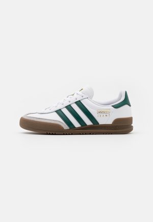 Witte Adidas sneaker met groene strepen, een gumkleurige zool en gouden "Jeans"-branding aan de zijkant, getoond in zijaanzicht tegen een witte achtergrond.