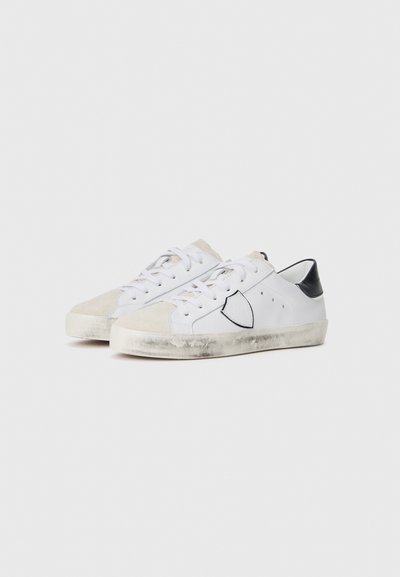 Białe sneakersy z zamszowym czubkiem, czarnym piętkiem, płaskim sznurowaniem i widocznymi śladami zużycia na podeszwach. Z boku znajduje się czarna obramowana detal z logo.